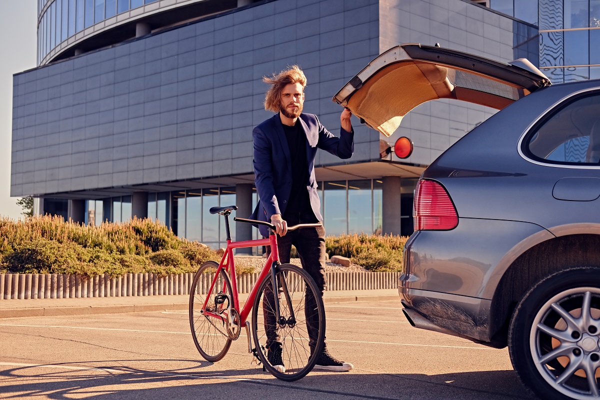 Bearded blond male with long hair puts fixed bicycle in the car's trunk.
